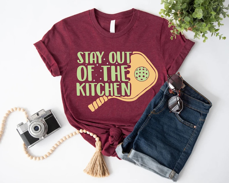 May include: A maroon t-shirt with the text "Stay out of the kitchen" and a yellow paddleball with a green ball. The t-shirt is paired with denim shorts and sunglasses.