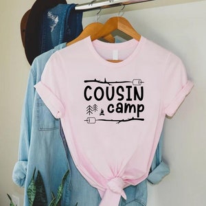 May include: Pink t-shirt with black text that reads "COUSIN camp" with a campfire and pine trees graphic.