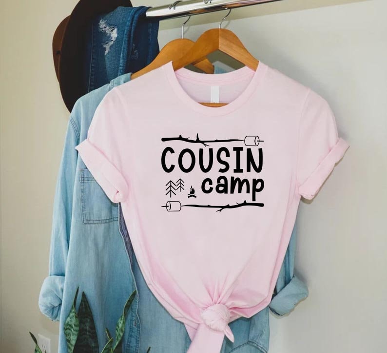 May include: Pink t-shirt with black text that reads "COUSIN camp" with a campfire and pine trees graphic.