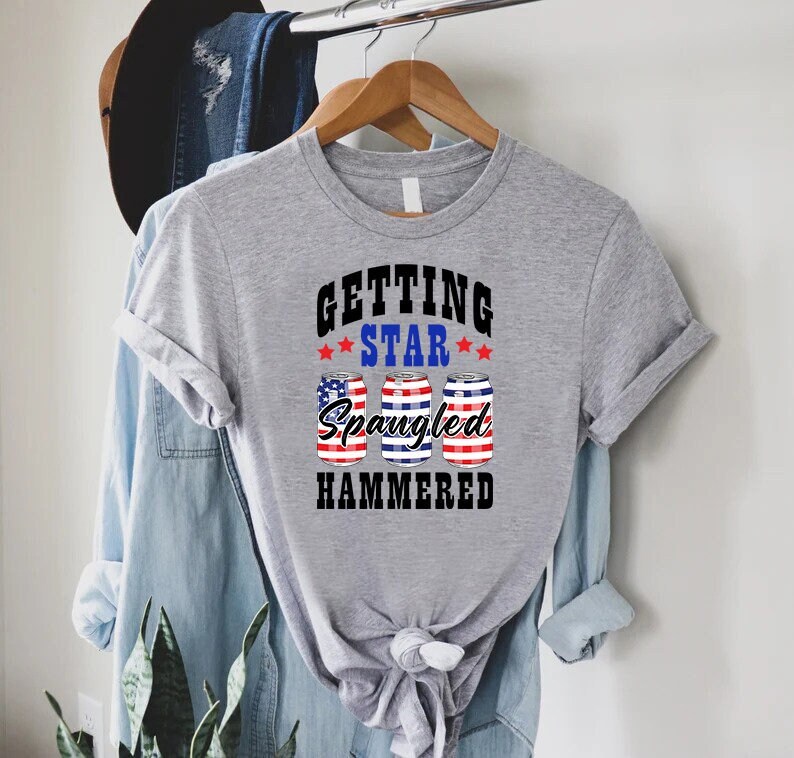 Op de afbeelding: Een grijs T-shirt met de tekst "Getting Star Spangled Hammered" in zwart. De tekst wordt omringd door drie rood-wit-blauwe blikjes met sterren.
