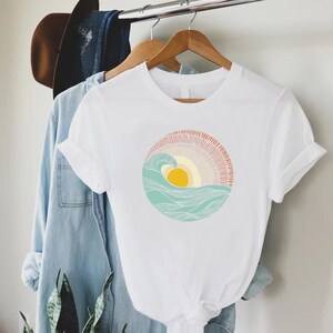 May include: White t-shirt with a graphic of a stylized sun setting behind a wave in a circle. The sun is yellow, the wave is blue and green, and the background is pink and orange.