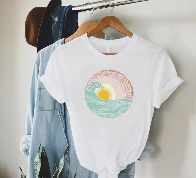 May include: White t-shirt with a graphic of a stylized sun setting behind a wave in a circle. The sun is yellow, the wave is blue and green, and the background is pink and orange.