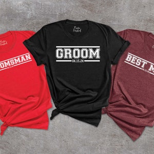 May include: Three t-shirts with a white text design on a black, red, and maroon background. The text on the shirts reads "Groom 04.15.24", "Groomsman 04.15.24", and "Best Man 04.15.24".