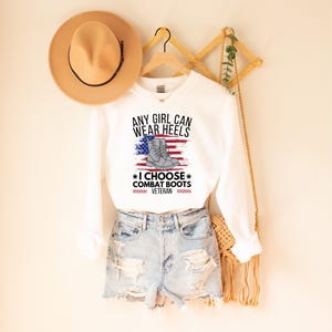 May include: White sweatshirt with the text "Any girl can wear heels *I choose combat boots* Veteran" and an American flag with a pair of combat boots on it.