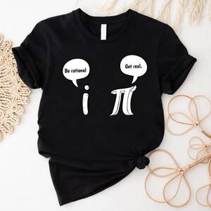 K&ouml;nnte beinhalten: Schwarzes T-Shirt mit einer wei&szlig;en Grafik der mathematischen Symbole 'i' und 'pi' mit Sprechblasen dar&uuml;ber. Die Sprechblase &uuml;ber 'i' sagt "Sei rational." und die Sprechblase &uuml;ber 'pi' sagt "Sei real."