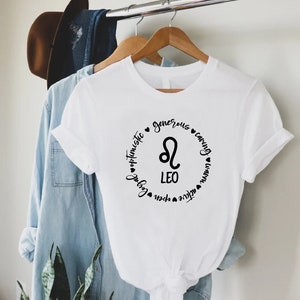 May include: White t-shirt with a black circle graphic featuring the Leo zodiac symbol and the words "Leo", "optimistic", "generous", "caring", "loyal", "open", "active", "warm", "fun", and "natural".