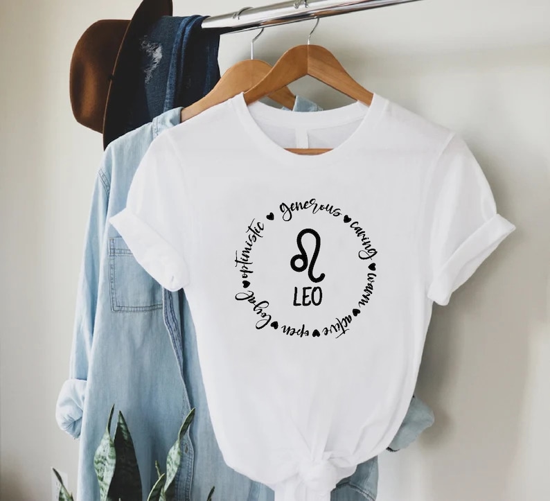May include: White t-shirt with a black circle graphic featuring the Leo zodiac symbol and the words "Leo", "optimistic", "generous", "caring", "loyal", "open", "active", "warm", "fun", and "natural".