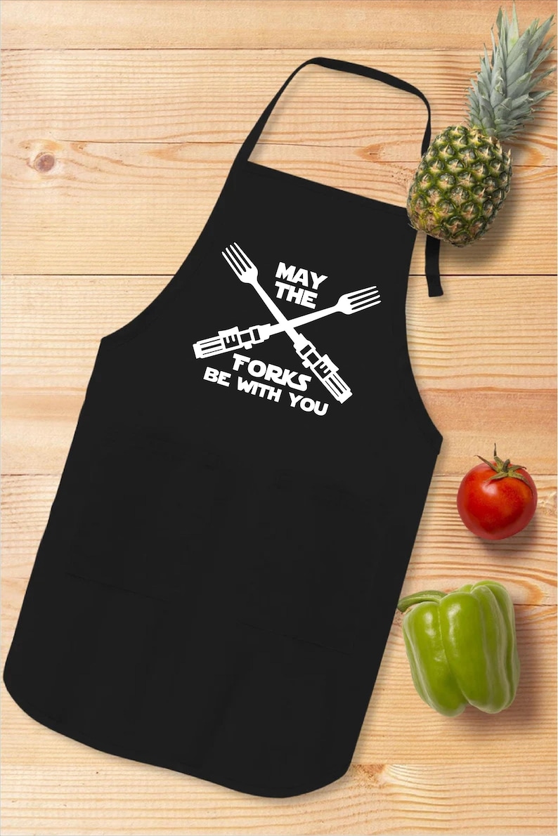 Pu&ograve; includere: Grembiule da cucina nero con un design bianco che dice "Che la forchetta sia con te" con due forchette incrociate che sembrano spade laser.