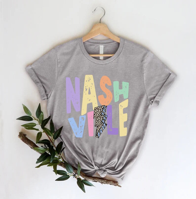 Puede incluir: Una camiseta gris con la palabra "NASHVILLE" impresa en letras de colores con un gr&aacute;fico de rayo en el centro.