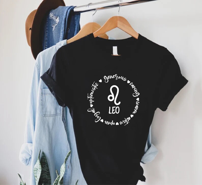 May include: Black t-shirt with a white circle graphic featuring the Leo zodiac symbol and the words "Leo", "optimistic", "generous", "caring", "loyal", "brave", "active", "fun", "passionate", and "warm".