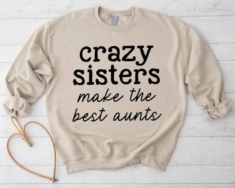 May include: A beige sweatshirt featuring the black text "crazy sisters make the best aunts". The long-sleeved sweatshirt has a crew neck. The text is in a bold, sans-serif font.