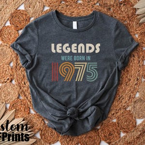 May include: A dark gray t-shirt with the text "Legends were born in 1975" in a retro font with colorful stripes.