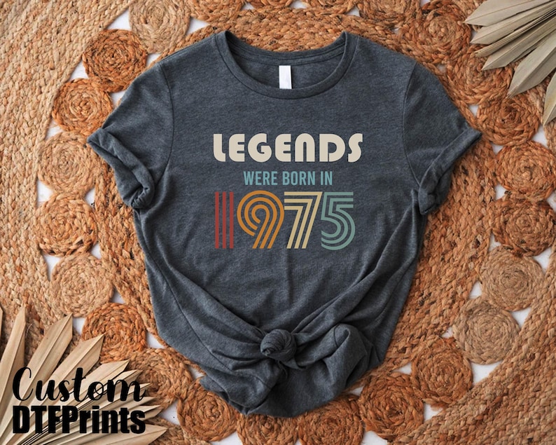 May include: A dark gray t-shirt with the text "Legends were born in 1975" in a retro font with colorful stripes.