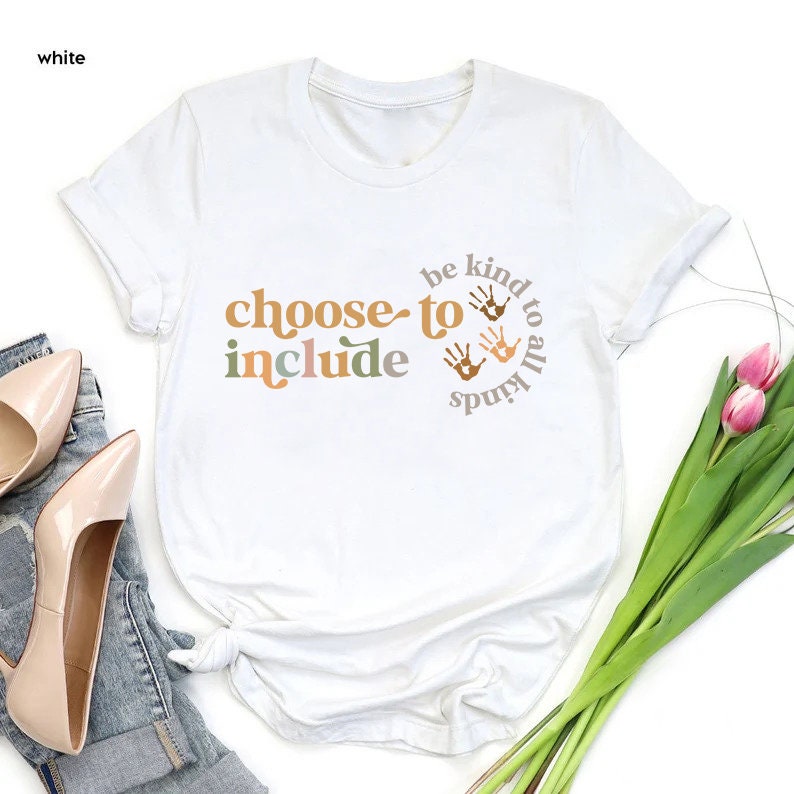 以下が含まれることがあります： オレンジ、茶色、緑色の文字で「choose to include」と書かれた白いTシャツ。また、手形のデザインと「be kind to all kinds」というフレーズが円形に配置されています。