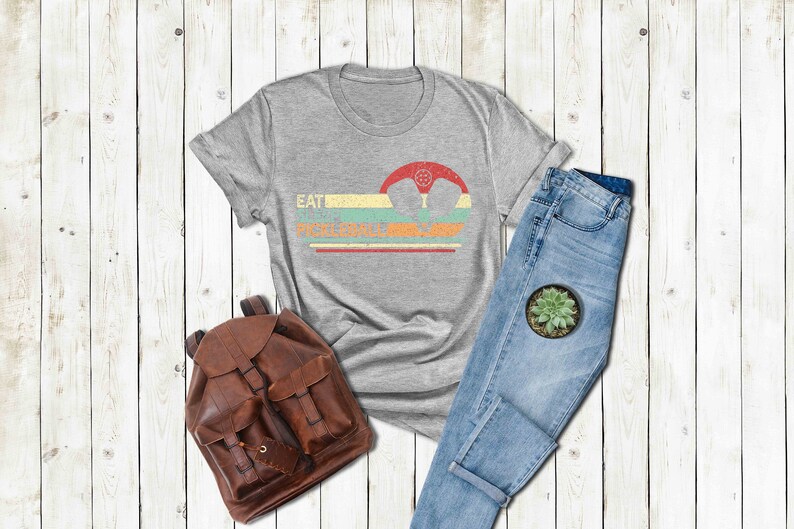 Pu&ograve; includere: Una maglietta grigia con una grafica in stile vintage che dice "Eat Sleep Pickleball Repeat". La grafica presenta una racchetta da pickleball e una palla in uno schema di colori retr&ograve;.