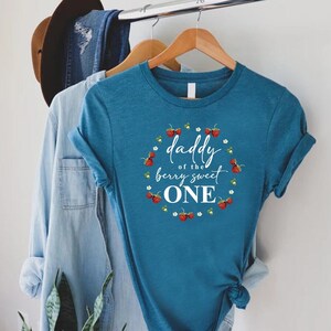 May include: A teal blue t-shirt with a white circle graphic that says "Daddy of the berry sweet one" surrounded by red strawberries and white flowers.
