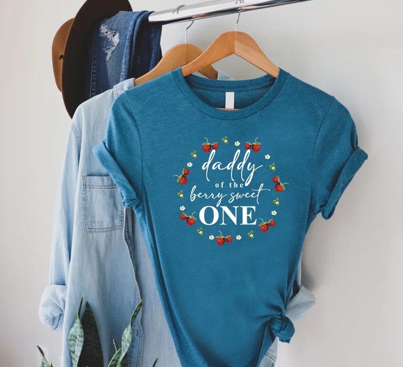 May include: A teal blue t-shirt with a white circle graphic that says "Daddy of the berry sweet one" surrounded by red strawberries and white flowers.