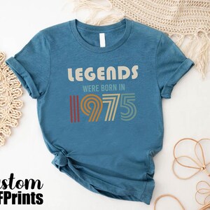 May include: A blue t-shirt with the text "Legends were born in 1975" in a vintage style font. The numbers are in a rainbow color scheme.