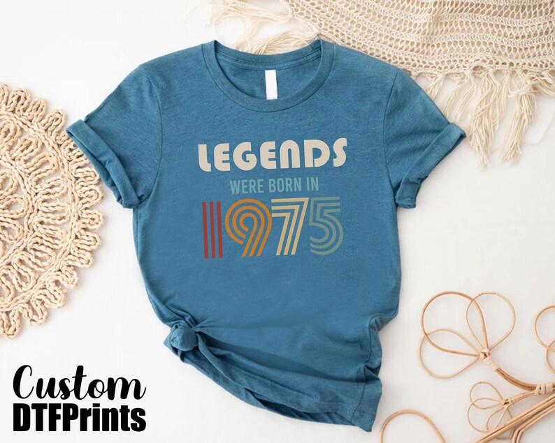 May include: A blue t-shirt with the text "Legends were born in 1975" in a vintage style font. The numbers are in a rainbow color scheme.