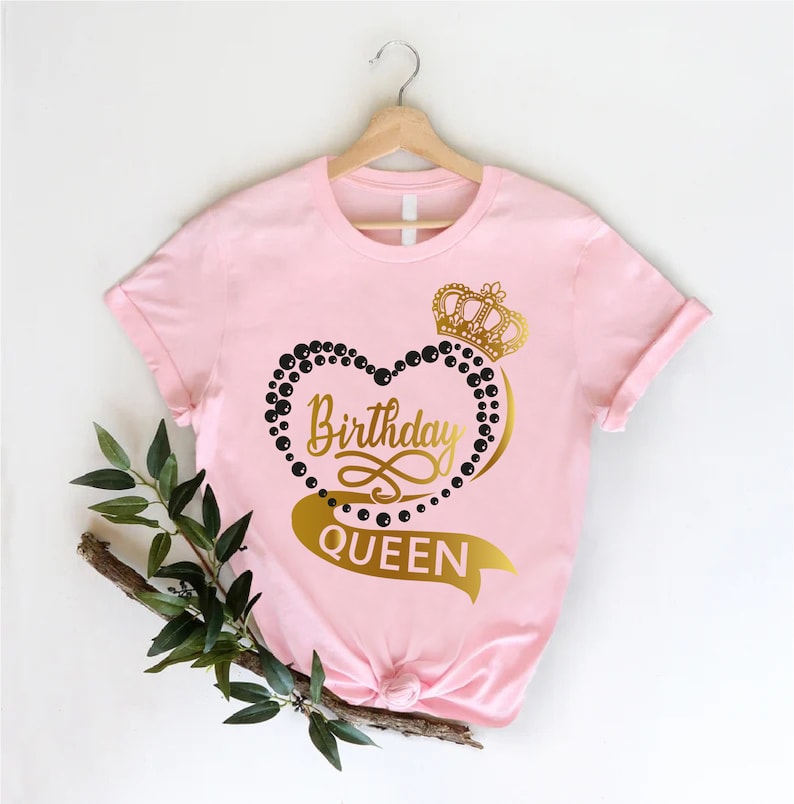 May include: Pink t-shirt with a gold heart design and the text "Birthday Queen". The heart is made of black beads and has a gold crown on top.