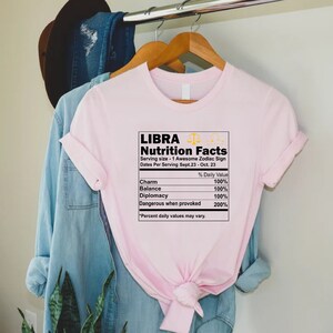 May include: Pink t-shirt with a graphic of a nutrition facts label for Libra, a zodiac sign. The label lists the daily value of charm, balance, diplomacy, and dangerous when provoked. The text reads "Libra Nutrition Facts Serving size - 1 Awesome Zodiac Sign Dates Per Serving Sept. 23 - Oct. 23 % Daily Value Charm 100% Balance 100% Diplomacy 100% Dangerous when provoked 200% *Percent daily values may vary."