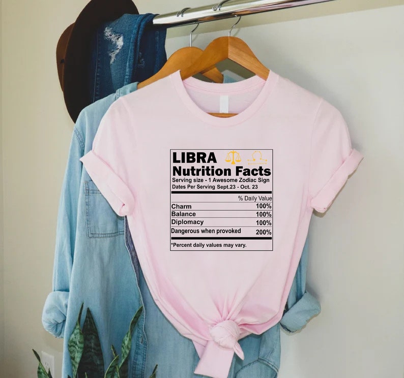May include: Pink t-shirt with a graphic of a nutrition facts label for Libra, a zodiac sign. The label lists the daily value of charm, balance, diplomacy, and dangerous when provoked. The text reads "Libra Nutrition Facts Serving size - 1 Awesome Zodiac Sign Dates Per Serving Sept. 23 - Oct. 23 % Daily Value Charm 100% Balance 100% Diplomacy 100% Dangerous when provoked 200% *Percent daily values may vary."