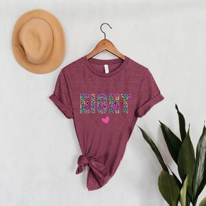 May include: A maroon t-shirt with the word "EIGHT" printed in rainbow leopard print. A small pink heart is below the word.