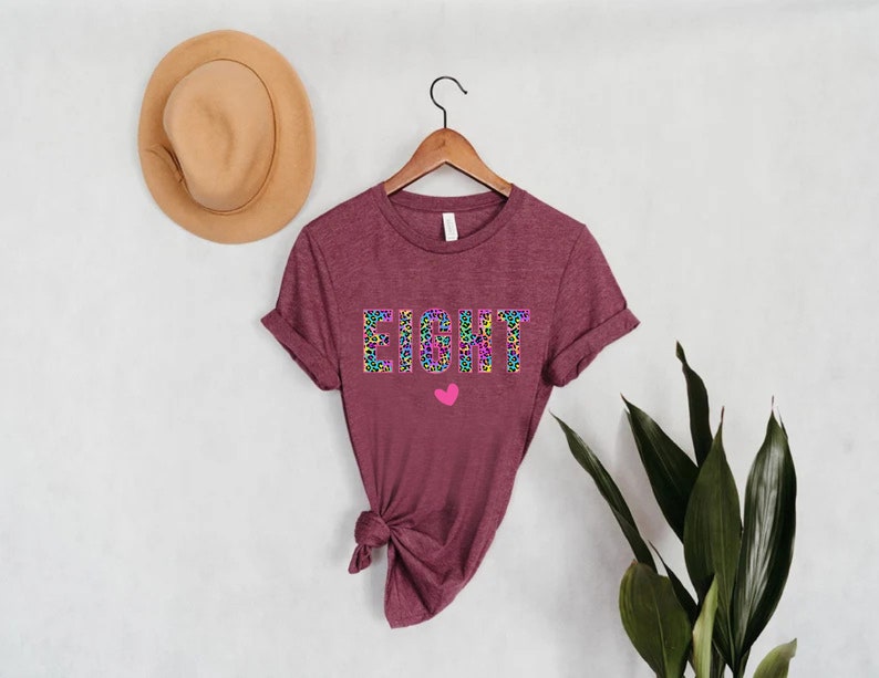 May include: A maroon t-shirt with the word "EIGHT" printed in rainbow leopard print. A small pink heart is below the word.