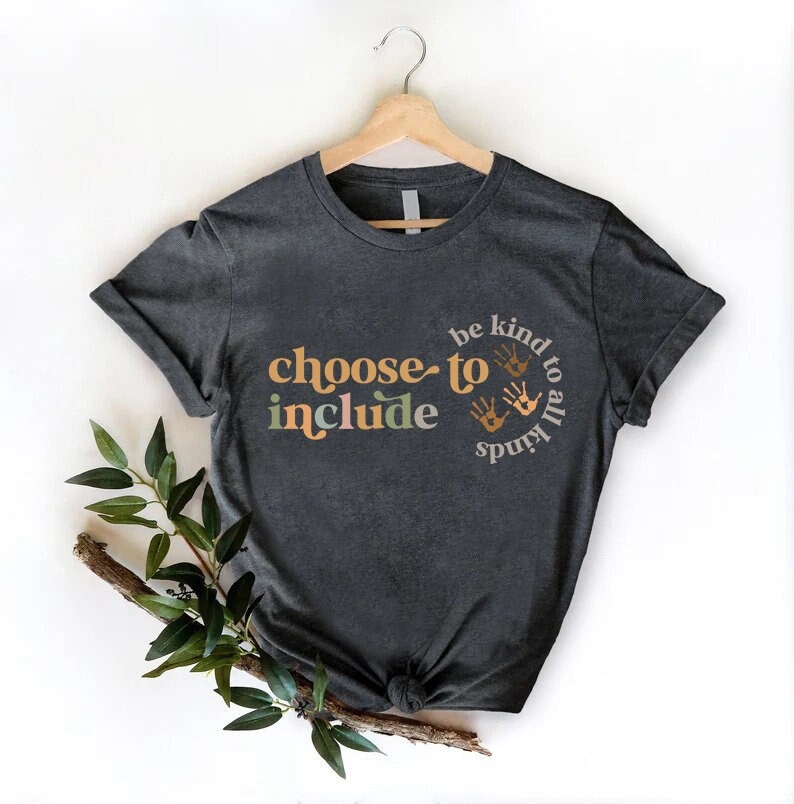 以下が含まれることがあります： オレンジと緑色の「choose to include」と、手形付きの「be kind to all kinds」という文字がプリントされた濃いグレーのTシャツ。木製のハンガーにかかっています。