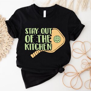 May include: Black t-shirt with a white print that says "Stay Out of the Kitchen" with a yellow pickleball paddle graphic.