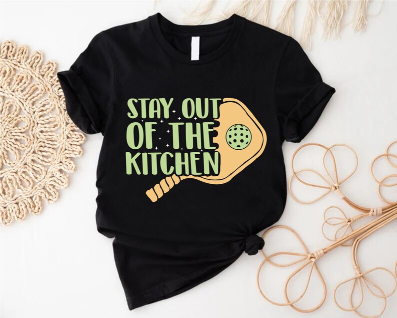 May include: Black t-shirt with a white print that says "Stay Out of the Kitchen" with a yellow pickleball paddle graphic.