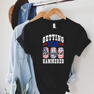 Op de afbeelding: Zwart T-shirt met een witte afbeelding met de tekst "Getting Star Spangled Hammered" met drie rood-wit-blauwe blikjes bier.