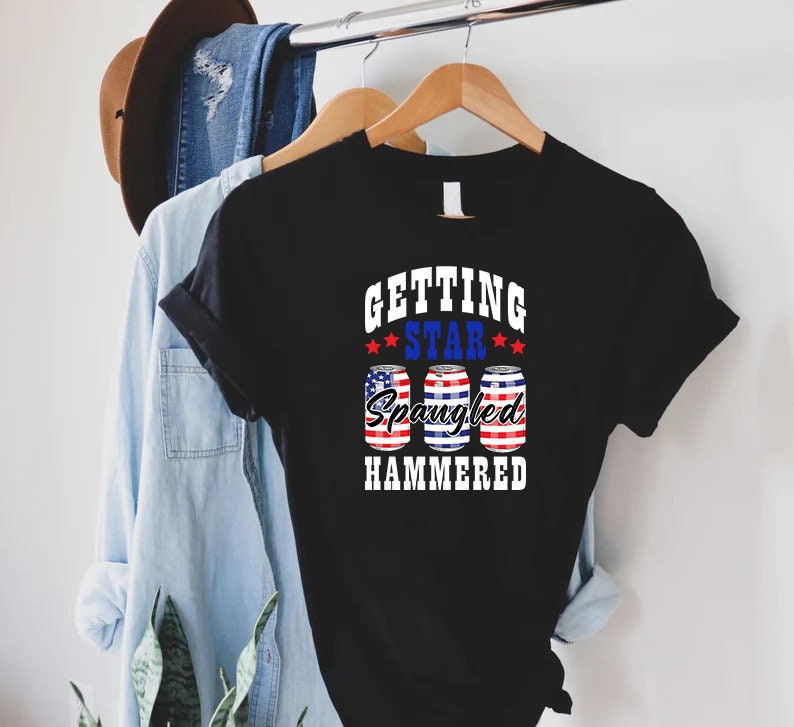 Op de afbeelding: Zwart T-shirt met een witte afbeelding met de tekst "Getting Star Spangled Hammered" met drie rood-wit-blauwe blikjes bier.