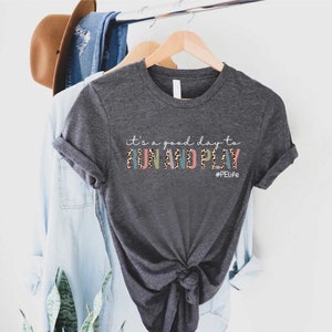 K&ouml;nnte beinhalten: Ein dunkelgraues T-Shirt mit dem Text "it's a good day to run and play" in bunten Buchstaben mit einem Leopardenmuster. Der Text "#PELife" ist unter dem Haupttext gedruckt.
