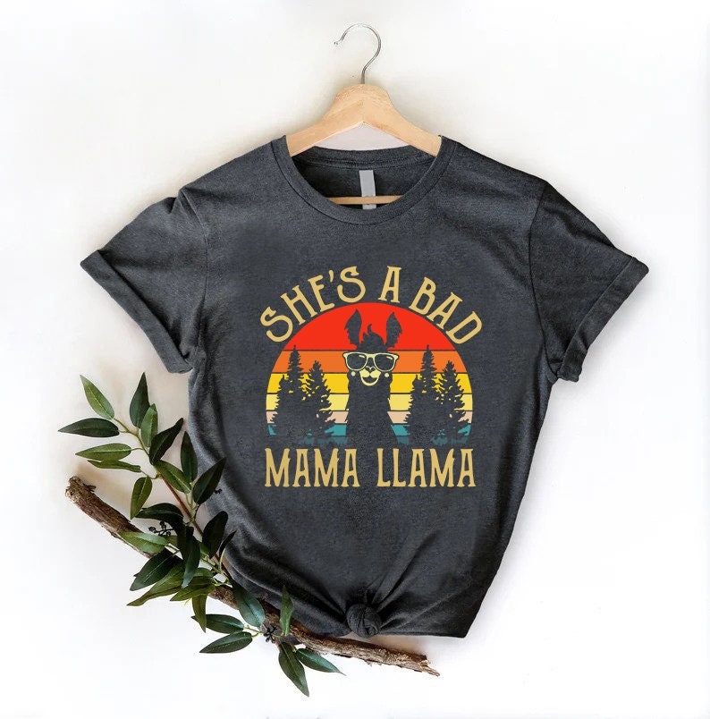 May include: A dark gray t-shirt with a vintage-style graphic of a llama wearing sunglasses in front of a sunset. The text "She's a Bad Mama Llama" is printed below the llama.