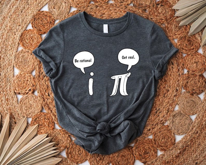 K&ouml;nnte beinhalten: Ein dunkelgraues T-Shirt mit einer wei&szlig;en Grafik der mathematischen Symbole 'i' und 'pi' mit Sprechblasen dar&uuml;ber. Die Sprechblase &uuml;ber 'i' sagt "Sei rational" und die Sprechblase &uuml;ber 'pi' sagt "Sei real."