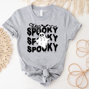 May include: A light gray t-shirt with the words "Stay Spooky" in black, dripping font, and a white ghost graphic. The shirt features a crew neck and rolled-up sleeves, perfect for Halloween.