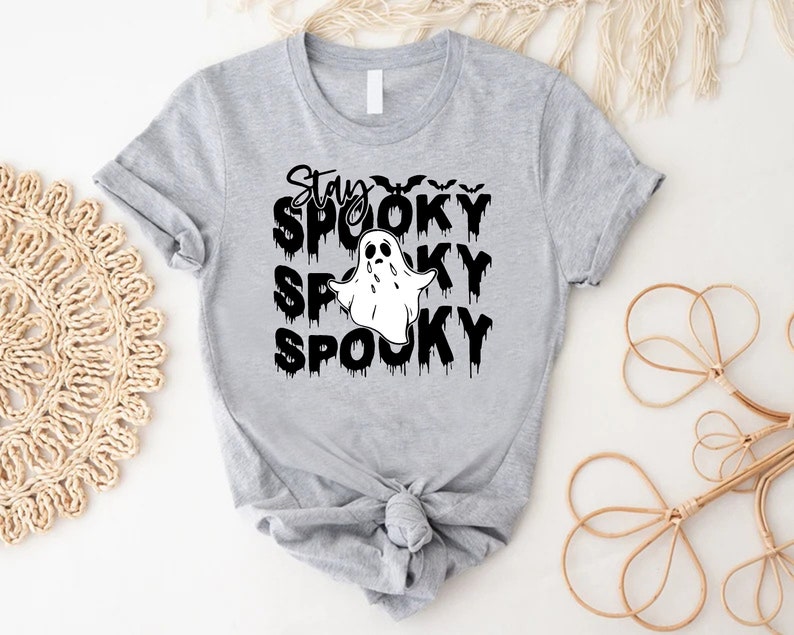 May include: A light gray t-shirt with the words "Stay Spooky" in black, dripping font, and a white ghost graphic. The shirt features a crew neck and rolled-up sleeves, perfect for Halloween.