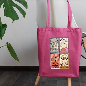 May include: Pink tote bag with a Halloween-themed design featuring four illustrated cards. The cards depict a ghost, a pumpkin, and a witch. The text on the cards reads "The Ghoul", "The Ghoul-ish", "The Black There", and "The Friendly Forest".