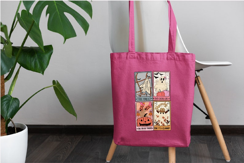 May include: Pink tote bag with a Halloween-themed design featuring four illustrated cards. The cards depict a ghost, a pumpkin, and a witch. The text on the cards reads "The Ghoul", "The Ghoul-ish", "The Black There", and "The Friendly Forest".