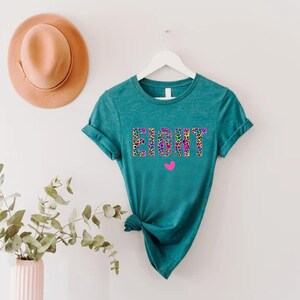 May include: Teal t-shirt with the word "EIGHT" printed in colourful leopard print letters. A small pink heart is below the word.