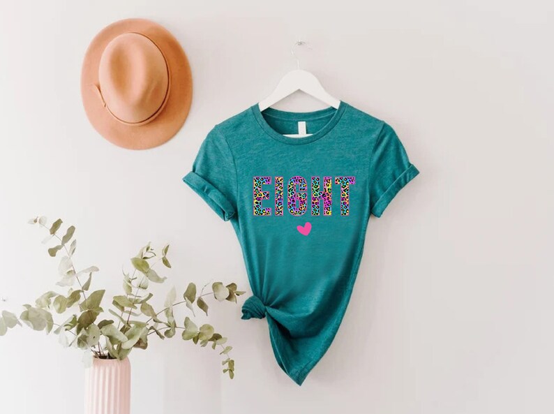 May include: Teal t-shirt with the word "EIGHT" printed in colourful leopard print letters. A small pink heart is below the word.