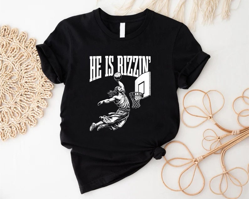 May include: Black t-shirt with a white graphic of a person dunking a basketball. The text "HE IS RIZZIN'" is above the graphic.
