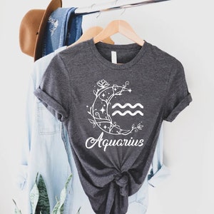 May include: A dark grey t-shirt with a white graphic of a crescent moon with a floral design and the zodiac symbol for Aquarius. The text "Aquarius" is written in a white cursive font below the moon.