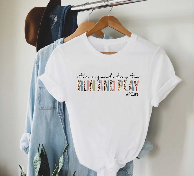 May include: White t-shirt with the text "it's a good day to RUN AND PLAY #PELife" printed on the front. The text is in a colorful font with a leopard print pattern.