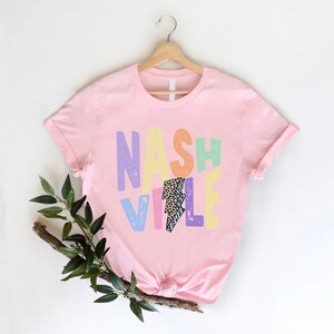 Puede incluir: Camiseta rosa con un gr&aacute;fico colorido que dice "Nashville" con un rayo en el medio.