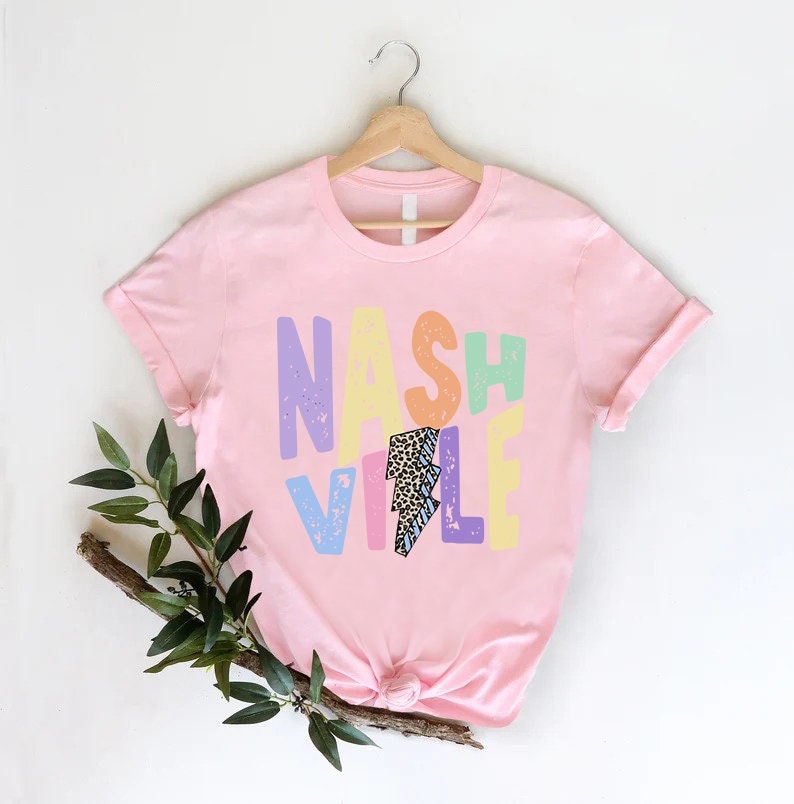 Puede incluir: Camiseta rosa con un gr&aacute;fico colorido que dice "Nashville" con un rayo en el medio.