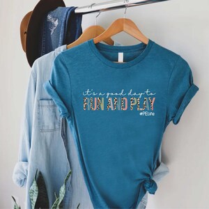 K&ouml;nnte beinhalten: Tealblaues T-Shirt mit dem Text "it's a good day to RUN AND PLAY #PELife" in bunten Buchstaben mit Leopardenmuster.