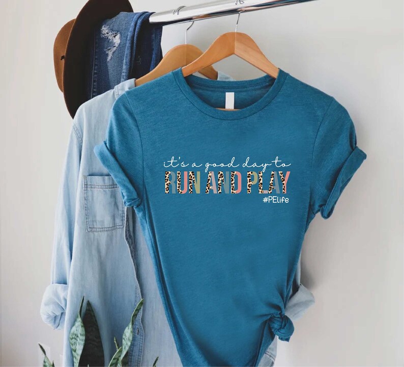 K&ouml;nnte beinhalten: Tealblaues T-Shirt mit dem Text "it's a good day to RUN AND PLAY #PELife" in bunten Buchstaben mit Leopardenmuster.