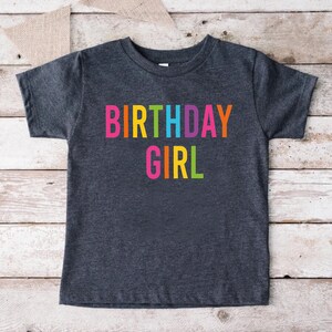 May include: A dark grey t-shirt with the words "BIRTHDAY GIRL" printed in rainbow colors.